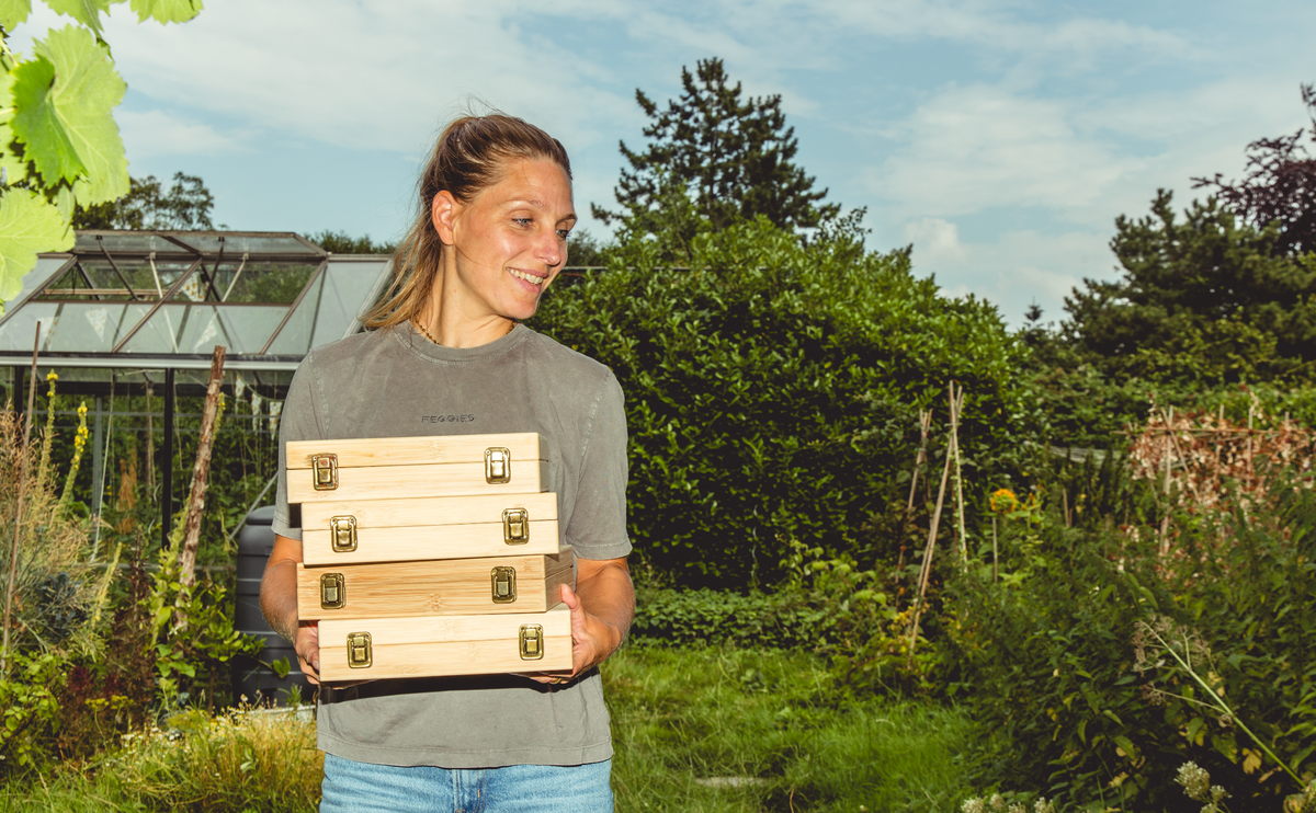 Kweker met een stapel bamboe zadenbewaarboxen in de moestuin, met een kas op de achtergrond